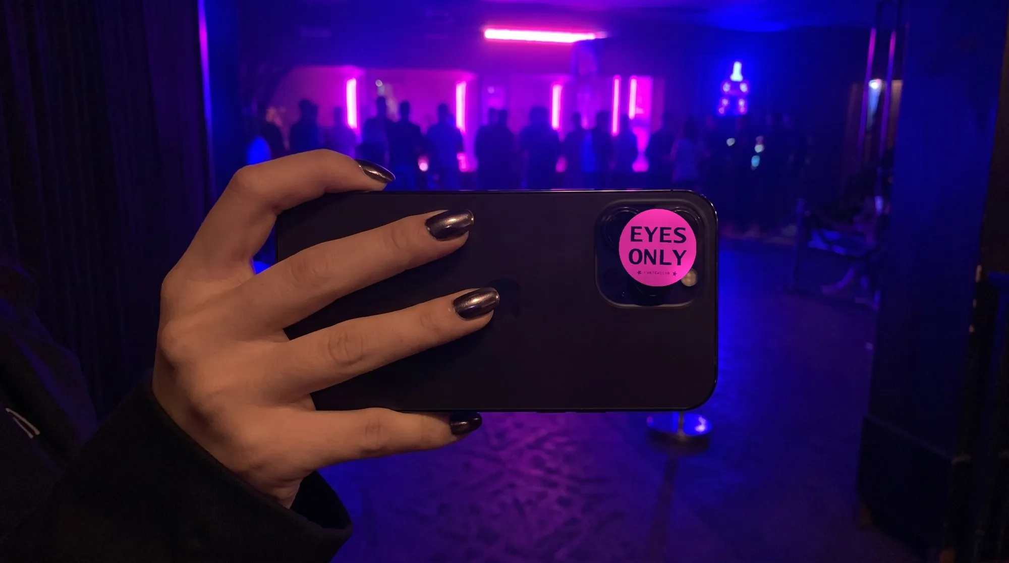 Il retro di un telefono nero tenuto in mano all'ingresso di un locale buio, con luce viola alle spalle. Sulle lenti della fotocamera è appiccicato un bollino rotondo magenta con la scritta «EYES ONLY» in viola scuro e, sotto, il marchio CURFEWCLUB fra due stelle.